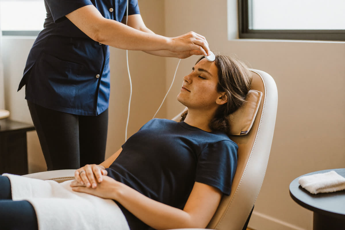 A practitioner places a small round scalp sensor at the hairline of a woman settling into a reclining chair in a bright studio room.