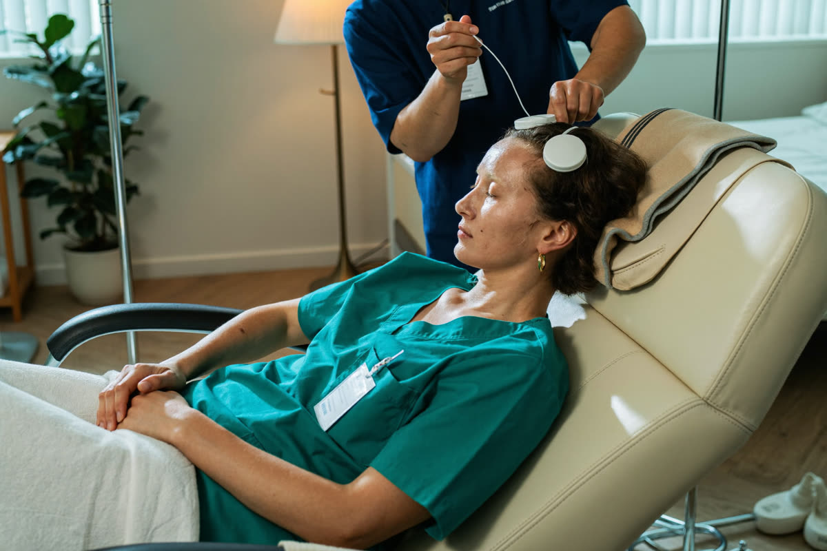 A nurse still in teal scrubs rests in a reclining studio chair in early morning light while a practitioner places a small scalp sensor.